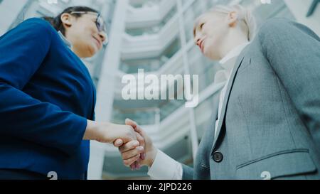 Low angle shot of businesswoman shaking hands and talking with female business colleague in suit in hall of modern office building indoors Stock Photo