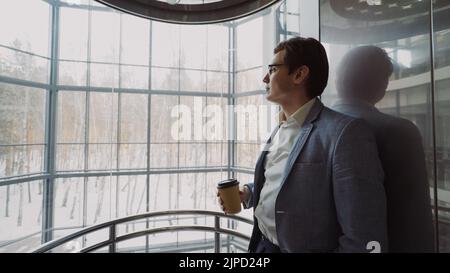 Young handsome businessman in suit drinking coffee moving down inside eleavator in modern business center indoors Stock Photo