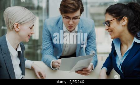 Close-up of cheerful businessman talking and discussing about financial report with business female colleagues sitting on sofa in meeting room indoors Stock Photo
