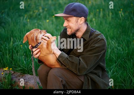 Dog as family member concept. Attractive caucasian man in baseball cap sitting on a grass in park and playing with small brown puppy biting him on finger. Young male with dachshund outdoors Stock Photo