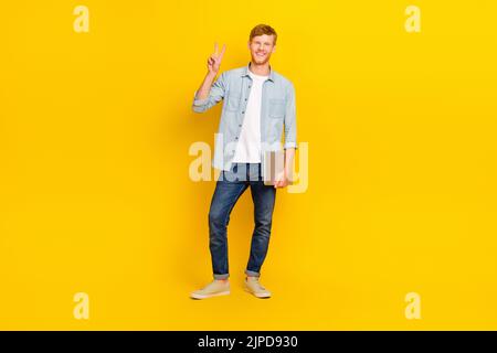 Full length photo of young happy programmer guy hold laptop showing v-sign friendly isolated on yellow color background Stock Photo