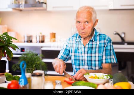 elderly man cuts vegetables for salad at the table in the kitchen Stock Photo