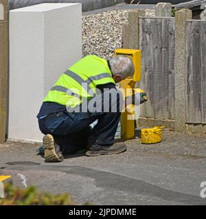 Workman from Essex Fire and Rescue Service sitting on pavement with paint pot painting yellow street hydrant sign wearing high visibility jacket UK Stock Photo