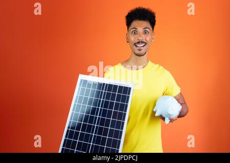 Handsome Black Man Holding a Piggy Bank and Solar Panel Isolated on Orange. Stock Photo