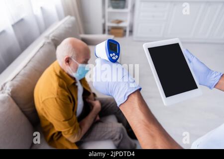 African american nurse holding pyrometer and digital tablet with blank screen near blurred pensioner in medical mask at home Stock Photo