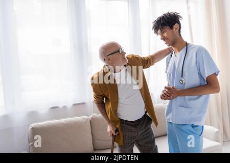 Smiling senior man with walking cane looking at african american nurse at home Stock Photo