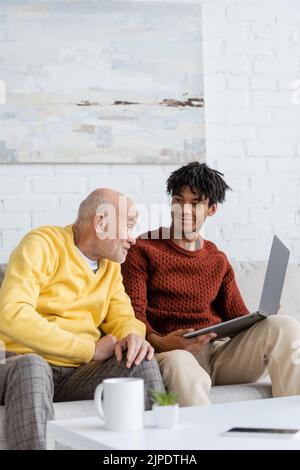 Senior man talking near smiling african american grandson with laptop at home Stock Photo