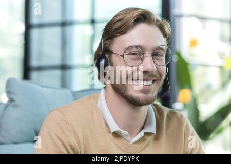Close-up photo of man wearing headset for video call, businessman working inside office building, smiling and looking at camera, employee at work wearing glasses Stock Photo
