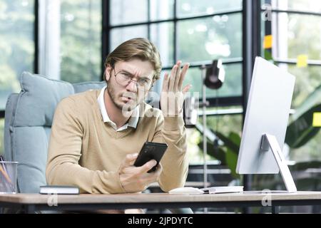 Dissatisfied businessman freelancer in glasses upset looking at camera man working inside office building holding phone and looking at camera with complaint Stock Photo