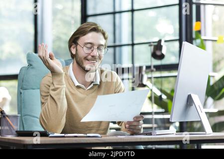 Happy blond man working inside an office building, young financier working with documents on paper work, businessman at computer smiling and reading financial report Stock Photo