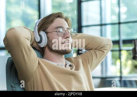 A man in an office on a chair with his arms folded is listening to music in headphones resting, a blond businessman is working inside an office building Stock Photo