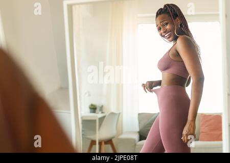 Fit black lady in front of mirror checking training and dieting results, looking at herself and smiling Stock Photo
