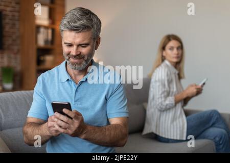 Happy adult caucasian husband with beard plays on smartphone, suffers from gadget addiction, ignores wife Stock Photo