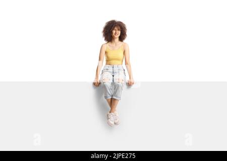 Full length portrait of a trendy young Caucasian female with afro hairstyle sitting on a blank panel isolated on white background Stock Photo