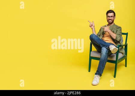 Positive middle aged man in casual sitting in armchair, pointing at copy space, advertising nice product, yellow background Stock Photo