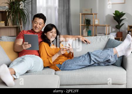 Relaxed asian couple chilling together at home, using gadgets Stock Photo