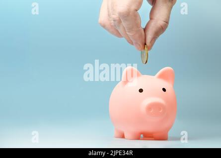Hand putting coin to pink piggy bank on a blue background. Copy space Stock Photo