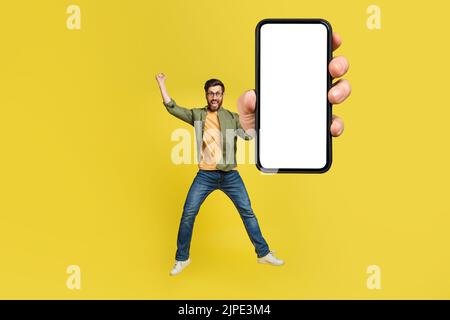 Full body length portrait of excited man jumping, making winner gesture and showing cellphone with empty space, mockup Stock Photo