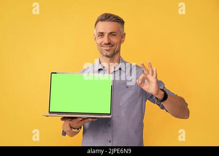 smiling man presenting computer showing ok gesture on yellow background, copy space, shopping. Stock Photo
