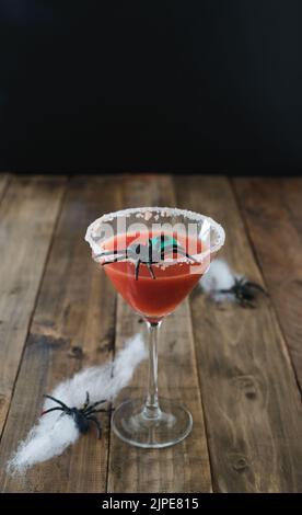 Terrifying red cocktail with spider on black background and wooden Halloween table with decoration. Vertical photo. Copy space. Stock Photo