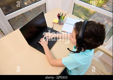 Top view of schoolboy wearing audio headset, typing text on laptop keyboard while doing homework at home. Homeschooling Stock Photo