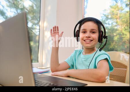 Adorable, smart schoolboy in headphones, waves with hand and smiles at the camera, while studying at an online school. Stock Photo