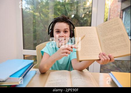 Smart student of online elementary school in headphones points at blurred text in textbook, answers teacher's questions Stock Photo