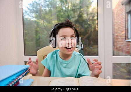 Cheerful schoolboy in headphones, smiles looking at camera while doing literature homework. Erudition. Back to school. Stock Photo