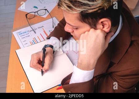 Top view of a man in a suit working at his desk full of graphs and writing in his notebook. Tired man working at his desk. Data analyst working hard. Stock Photo