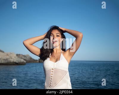 Self assured young Hispanic female tourist in white dress with long dark hair smiling and touching head while looking at camera on wavy seashore under Stock Photo