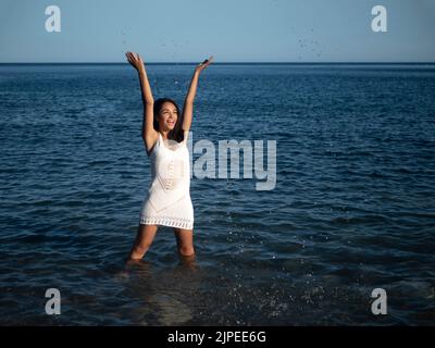 Cheerful young ethnic female tourist with long dark hair in crochet dress smiling brightly and looking away while standing in shallow sea and splashin Stock Photo