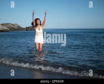 Full body of excited young Hispanic female traveler with long wavy hair in white summer dress walking in sea and splashing water while having fun duri Stock Photo
