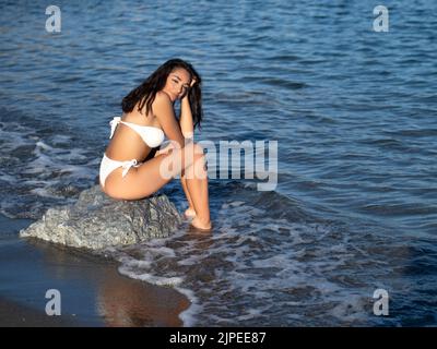Side view of content young Hispanic lady with long dark hair in white bikini relaxing on boulder on wet sandy beach and smiling while enjoying sunny d Stock Photo