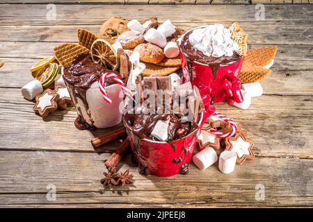 Christmas styled overloaded hot chocolate mugs with various toppings – chocolate slices, spices, sweets, cookies, candy and gingerbreads, wooden cozy Stock Photo