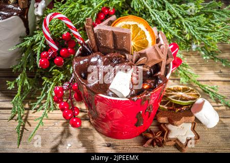 Christmas styled overloaded hot chocolate mugs with various toppings – chocolate slices, spices, sweets, cookies, candy and gingerbreads, wooden cozy Stock Photo