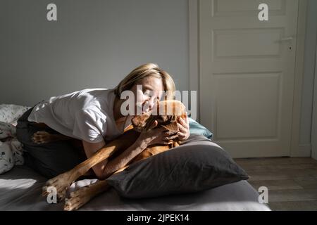 Lonely mature blonde woman enjoys hugging sleepy Vizsla dog lying on pillow in morning Stock Photo