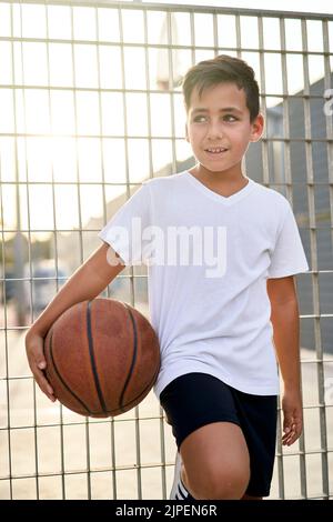 A cute young boy plays basketball on the street playground in summer. Teenager in a white t-shirt with orange basketball ball outside. Hobby, active l Stock Photo