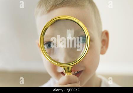 Faces of a boy holding a magnifying glass in his hand and looking cheerfully into the camera through it. Close-up. Stock Photo