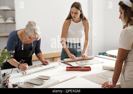 Pottery workshop in studio. People working with clay on the table. Adults learning to do ceramic plates. Pottery as hobby and leisure activity Stock Photo
