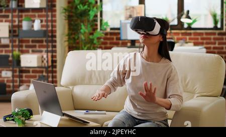 Playful woman wearing virtual reality headset scrolling through metaverse cyberspace while working from home. Asian person wearing futuristic VR goggles while interacting with cyber reality. Stock Photo