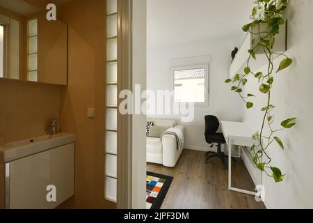 Simple bathroom with a small vanity unit with white porcelain sink and frameless mirror and access to a living room with windows, plants and a desk wi Stock Photo