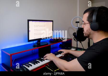 caucasian latin young man, with black clothes, headphones, beard and glasses, studying and practicing electric piano at home by online classes, sittin Stock Photo