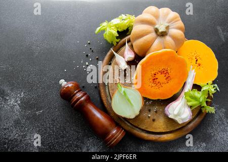 Fresh bright vegetables are laid out on a wooden cutting board: butternut squash, green celery leaves. Black uneven background, copy space. Stock Photo