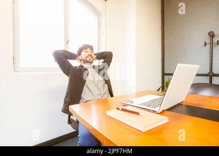Businessman sitting at the head of the meeting table with his hands behind his head, relaxed and happy to have finished his work Stock Photo