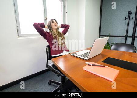 Businesswoman sitting at the head of the meeting table with his hands behind his head, relaxed and happy to have finished his work Stock Photo