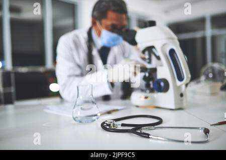Where medical solutions are made. a stethoscope on a laboratory table with a scientist using a microscope in the background. Stock Photo