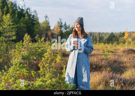 Young woman in hipster hat and denim coat walks past autumn trees looking at sunset Stock Photo