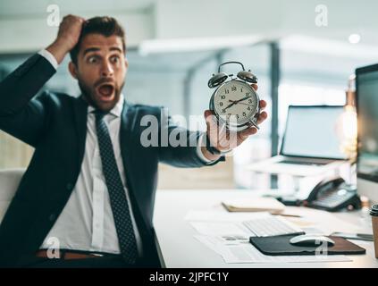 Oh dear is that the time already. a surprised young businessman holding an alarm clock while being seated in the office. Stock Photo