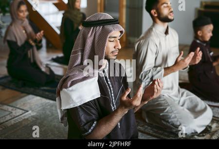 Young muslim family pray together, sit on knees at home practicing Islam. Religious women wearing hijab and spiritual men wear traditional attire Stock Photo