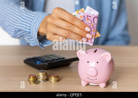 Close-up of a woman's hands put euro paper money in a piggy bank, next to a calculator on the table. economy, crisis and inflation Stock Photo